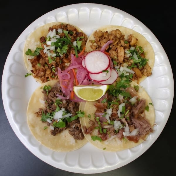 Four street tacos with various meats, cilantro, onions, pickled radishes, and a lime wedge on a white plate.
