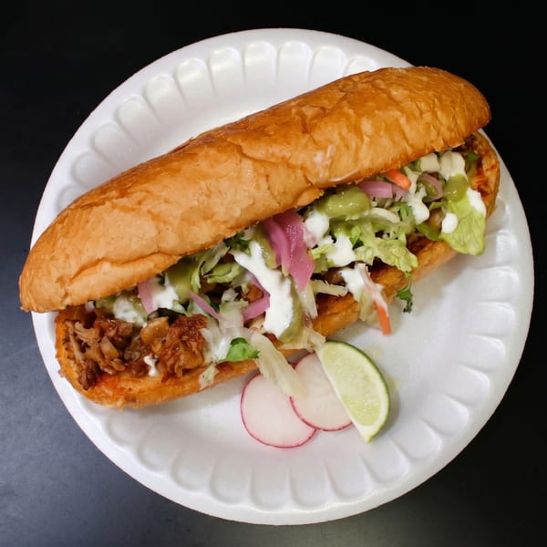 A large Mexican torta sandwich filled with meat, lettuce, pickled onions, and sauces, served with radish and lime.