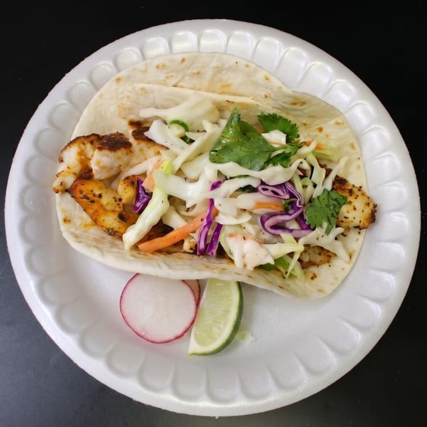 A fish taco on a white plate, topped with cabbage slaw, cilantro, a radish slice, and lime.