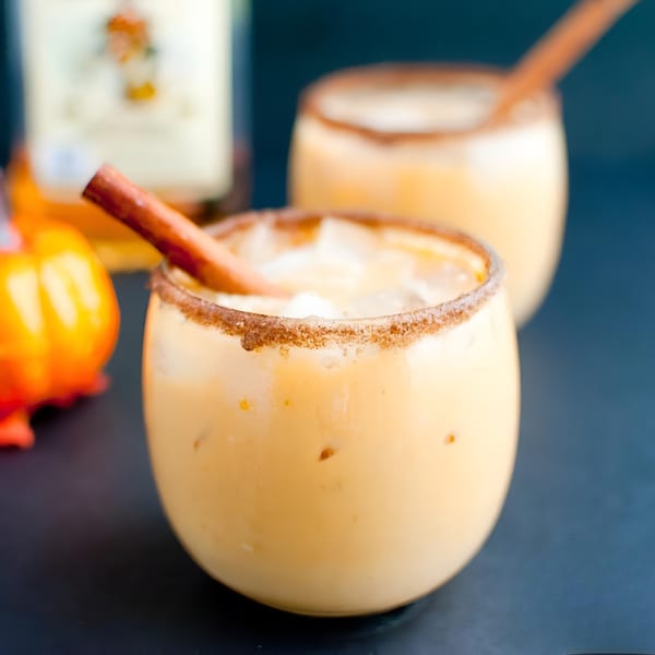 Two pumpkin spice cocktails with cinnamon sticks and sugar-rimmed glasses, with a small pumpkin and liquor bottle.