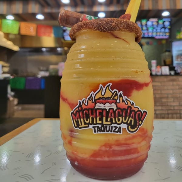 A large Michelaguas! Taquiza mango drink with chili powder rim and a small hot sauce bottle.